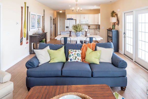 Bright open-plan living room with navy sofa, coastal artwork, and white kitchen visible beyond.