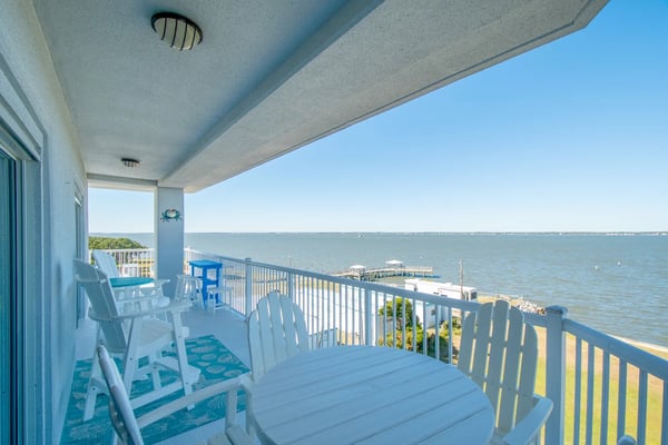 Covered waterfront balcony with white Adirondack chairs and round table looking out over the sound, with a boat dock and open water beyond